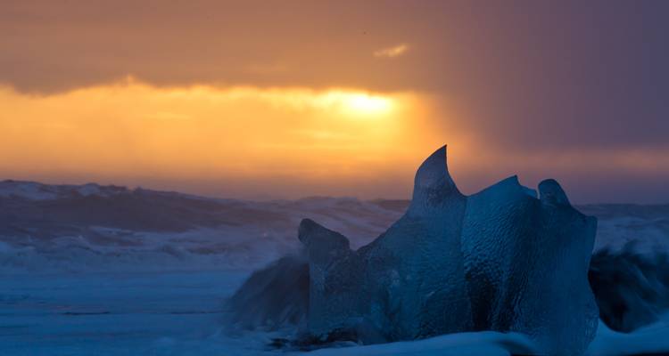 Gloeiende zonsopgang achter kolkende wintergolven en kristallijne ijsblokken op een zwart strand