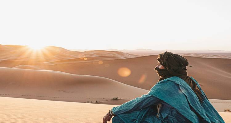 Un nomade vêtu de robes bleu-vert est assis sur une dune et contemple le lever du soleil sur les vagues infinies du désert.