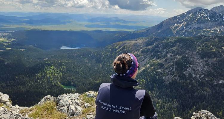 Wanderer sitzt auf felsigem Vorsprung und bewundert das weitläufige Alpenpanorama mit Wäldern, Seen und Gipfeln