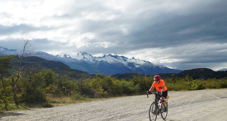 Cyclist in bright jersey riding gravel road with snow-capped Andes under dramatic clouds