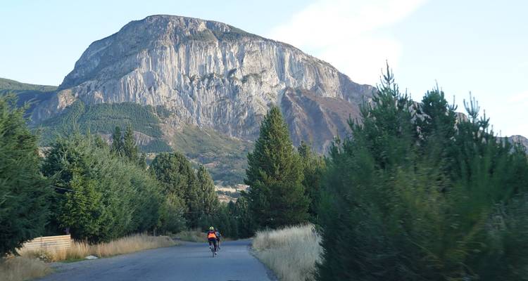 Lone cyclist approaching a dramatic sheer cliff mountain through pine-lined road