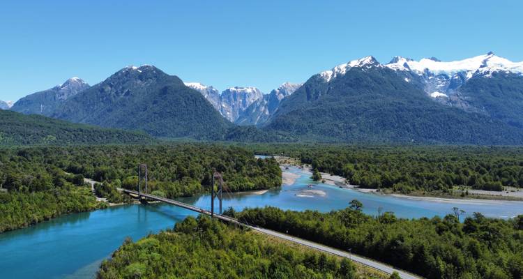 Aerial view of a suspension bridge crossing a turquoise river amid dense forest with dramatic snow-capped mountains in the background.