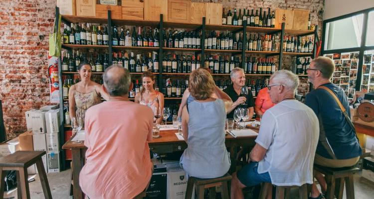 Small group seated at a rustic table inside a brick-walled wine shop enjoying a tasting with shelves of bottles behind them.