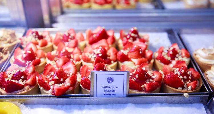 Tray of vibrant strawberry tartlets labelled in Portuguese on a bakery counter.