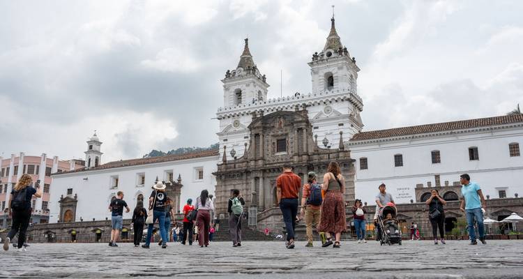 Los turistas pasean por la plaza empedrada frente a la histórica Iglesia Blanca de San Francisco de Quito bajo un cielo nublado.
