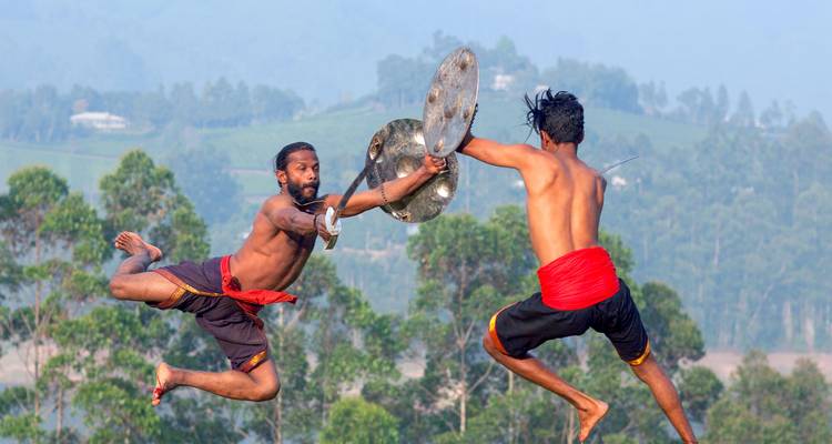 Two martial artists leap mid-air wielding shields in a dramatic Kerala landscape.