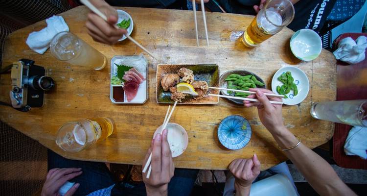 Des mains avec des baguettes se tendent vers des assiettes partagées de plats d'izakaya et des bières sur une table en bois.