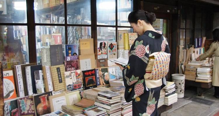 Une femme en kimono à motifs floraux parcourt des piles de livres anciens dans une boutique japonaise calme.