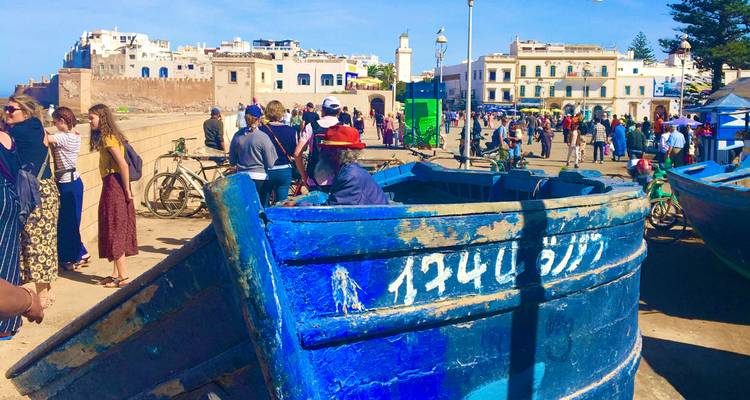 Druk havenplein in Essaouira met veel volk, fietsen en een groot blauw visserbootje op de voorgrond.