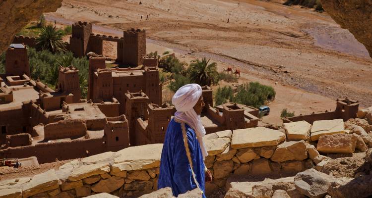 Een Berber-man in blauwe robe kijkt uit over de klei kasbah's en droge riverbedding van Ait Benhaddou.