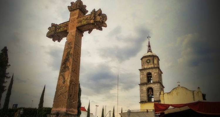 Un monumento de cruz de piedra se alza frente a un campanario de iglesia histórica bajo cielos nublados.