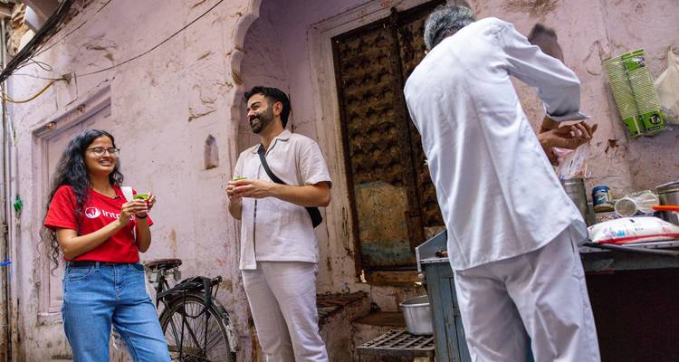 Reizigers chatten en nippen aan chai in een smal, pastelgekleurd steegje terwijl een lokale theeverkoopman drankjes bereidt naast een fiets.