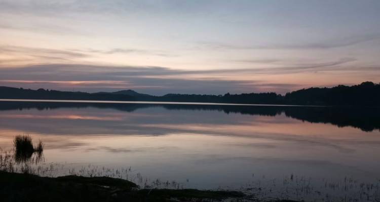 Las aguas tranquilas del lago reflejan los tonos pastel suaves del cielo justo después de la puesta de sol con colinas lejanas en silueta.
