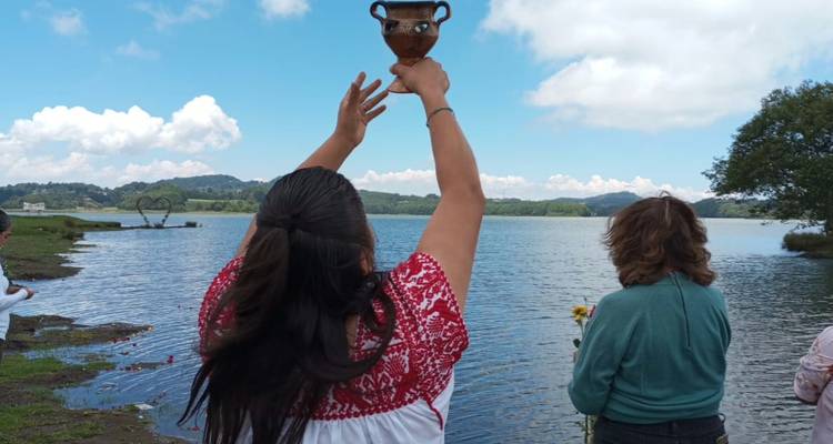 Dos mujeres realizan un ritual a orillas de un lago, una levanta una vasija decorada hacia el cielo mientras flores flotan en el agua.