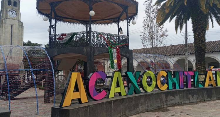 Letras 3-D coloridas que forman la palabra ACAXOCHITLÁN se encuentran frente a una glorieta de hierro forjado decorada con banderas mexicanas en una plaza del pueblo.