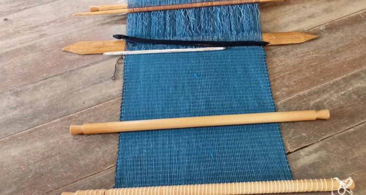 Las herramientas de tejido y un textil azul a medio terminar están dispuestos en tablones de madera.