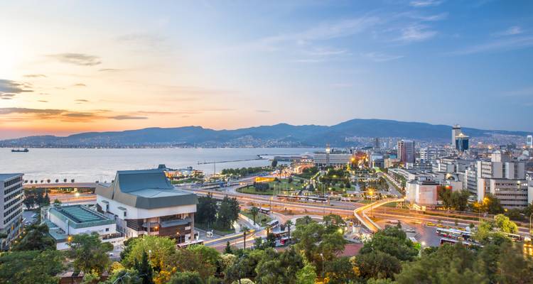 Vue surélevée du coucher de soleil sur l'horizon d'une ville côtière avec une baie sinueuse, des routes animées et des montagnes lointaines.