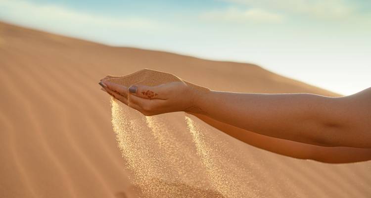 A hand lets fine desert sand stream through the fingers against a dune backdrop.
