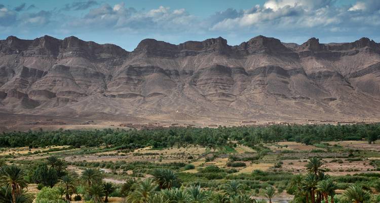 Sharp rocky escarpment towers over a lush palm-filled valley and patchwork farms in southern Morocco.