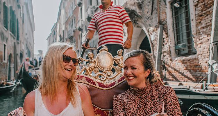 Twee vrouwen lachen op een versierde gondel terwijl de gondelier door een Venetiaans kanaal stuurt.