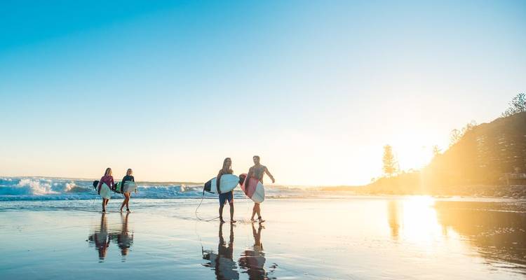 Grupo de surfistas llevando tablas por una playa reflectante al amanecer con luz dorada sobre las olas.