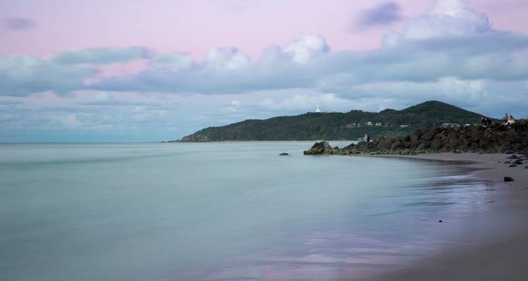 Escena tranquila del atardecer en tonos pastel suaves del océano tranquilo con un promontorio distante y un faro bajo cielos rosas y azules.