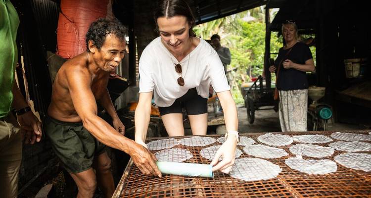 Visitors help a local craftsman roll rice paper sheets on bamboo racks in a traditional workshop.