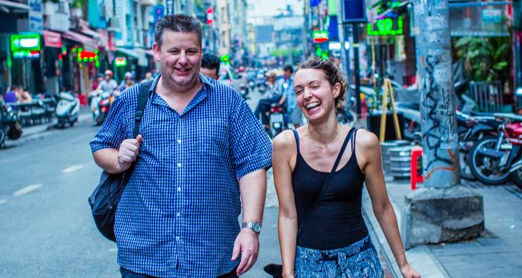 Ein lächelnder Mann und eine Frau spazieren eine lebhafte Stadtstraße hinunter, die mit Rollern und Leuchtreklamen gefüllt ist.