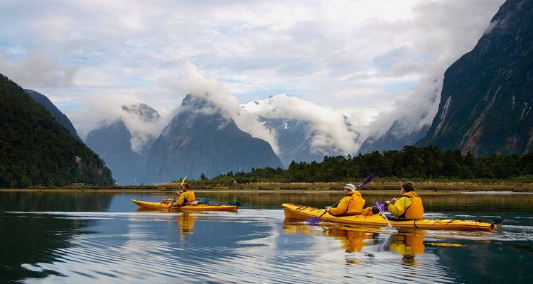 Kayakers paddle on mirror-calm water amid dramatic cloud-cloaked peaks of Milford Sound