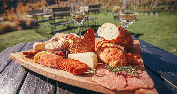 Rustic wooden board loaded with cheeses, meats and bread with wine glasses in a sunny vineyard setting