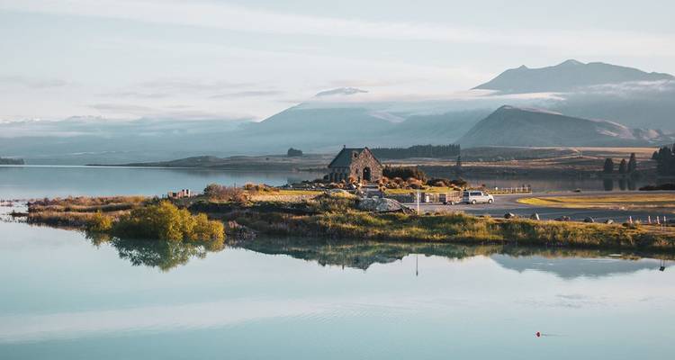 Stone church on a peninsula beside a calm turquoise lake with distant mountains under soft light