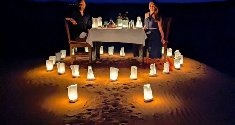 Mise en place pour un dîner romantique aux chandelles pour deux personnes sur le sable du désert la nuit, entourée de sacs de lanternes lumineuses.