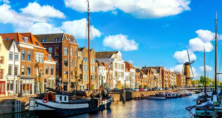 Scène de canal ensoleillée avec des maisons de ville traditionnelles hollandaises, des bateaux amarrés et un moulin à vent sous un ciel bleu avec des nuages épars.