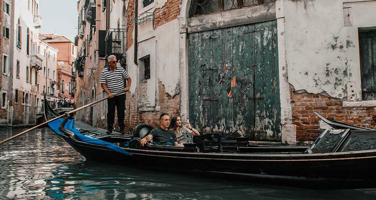 Un gondolier dirigeant une gondole avec des touristes assis dans un canal vénitien étroit bordé de bâtiments usés par le temps.
