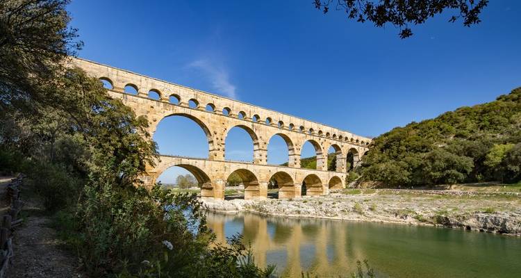 L'aqueduc romain historique du Pont du Gard s'étend sur une rivière calme sous un ciel bleu éclatant avec une verdure environnante.