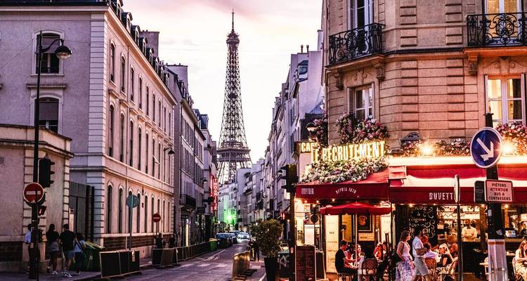 Scène de rue parisienne au crépuscule avec la Tour Eiffel encadrée par des bâtiments classiques et une terrasse de café animée remplie de clients.