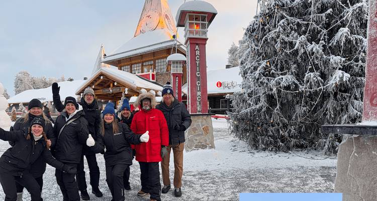 Des voyageurs souriants posent pour une photo devant le marqueur du Cercle arctique dans le village du Père Noël enneigé.