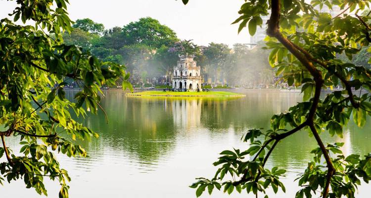 Turtle Tower op een klein eilandje in Hanoi's Hoan Kiem Lake, omlijst door bladrijke takken en ochtendmist.