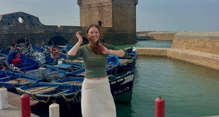 Mujer sonriente posando junto a barcas de pesca azules y murallas de piedra del puerto de Essaouira.
