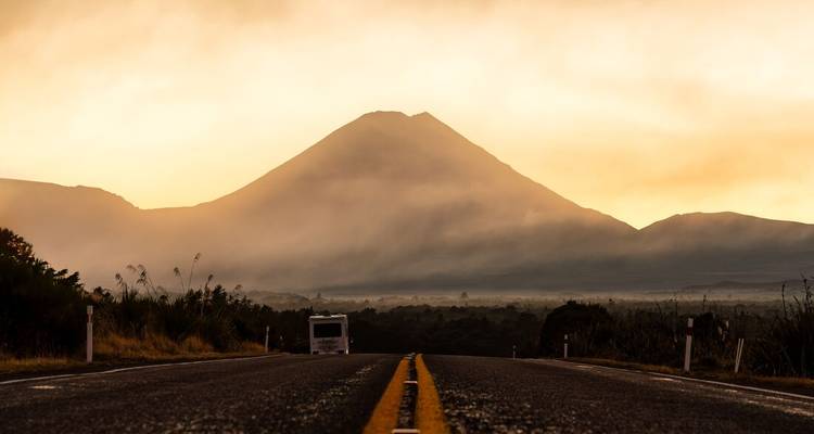 Dramatische Aussicht auf einen Bus, der bei Sonnenaufgang auf einen Kegelvulkan zufährt, mit nebligen Ebenen rund um Tongariro.