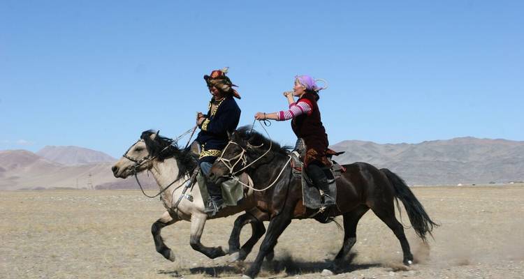 Zwei mongolische Reiter reiten Pferde über eine offene Ebene mit entfernten kahlen Bergen.