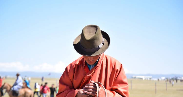 Porträt eines mongolischen Mannes in traditionellem Deel und Filzhut, der auf ein Steppenfeld hinabblickt.