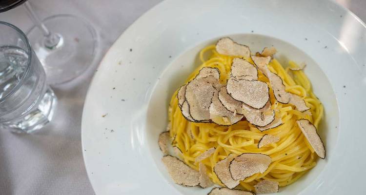 Un bol blanc de pâtes fraîches est généreusement garni de fines tranches de truffe à côté d'un verre de vin.