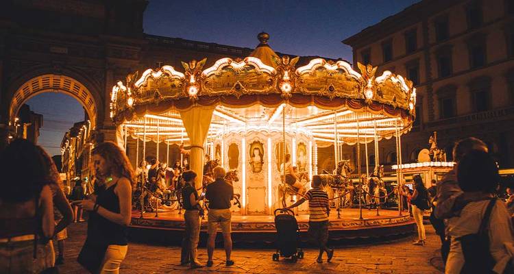 Un carrousel orné tourne brillamment sur une place la nuit tandis que les gens se rassemblent autour sous des lumières chaleureuses.