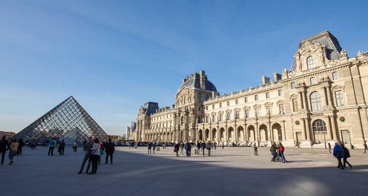 Large place du Louvre avec sa pyramide de verre et ses ailes de palais historiques sous un ciel bleu clair, des visiteurs dispersés ça et là.