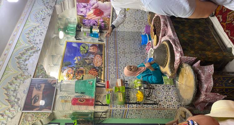 Local woman in a tiled stall prepares food surrounded by jars of pickles and colorful wall art in a Moroccan market.
