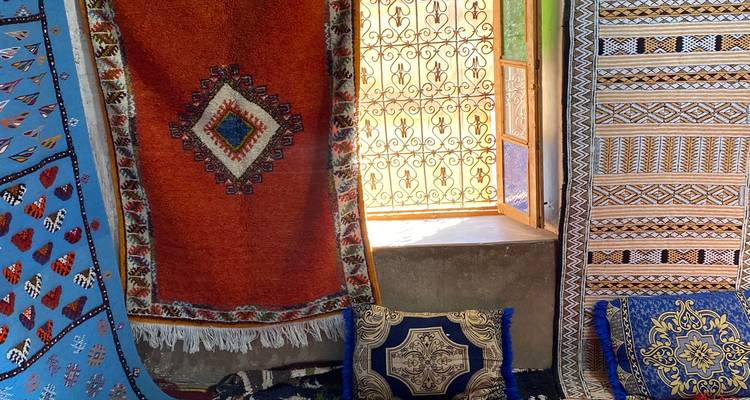 Sunlit display of colorful Moroccan carpets and cushions beside a wrought-iron window.