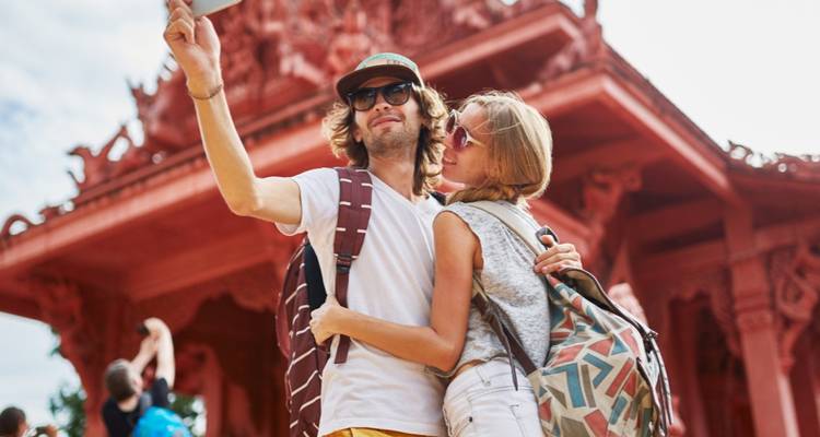 Un jeune couple de routards s'étreint et prend un selfie devant un temple rouge orné.