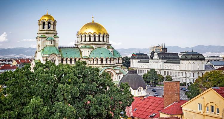 Les dômes dorés de la cathédrale Alexandre Nevski s'élèvent au-dessus des arbres feuillus et des toits colorés de Sofia avec les montagnes lointaines.