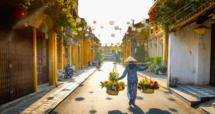 Vietnamese vendor in conical hat carrying flower baskets down a lantern-strung yellow street at sunrise.